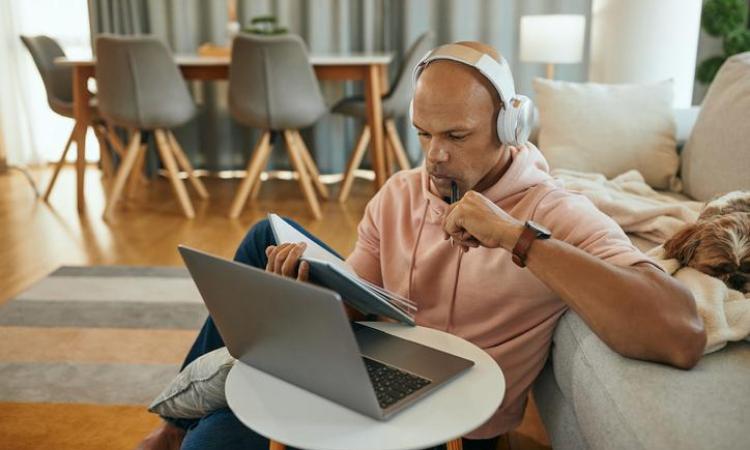 A man wearing headphones and holding a notebook looks at a laptop computer.