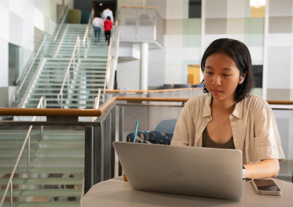 Female student working on a laptop at a table in a modern campus building with a glass staircase in the background.
